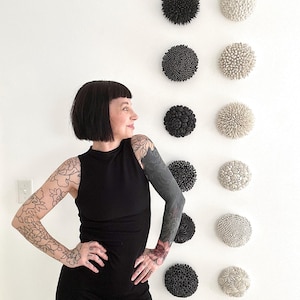 May include: A collection of decorative wall art pieces in various textures and colors, including black and off-white. The pieces are arranged in a grid pattern on a white wall. The art pieces have a round shape and a textured surface.