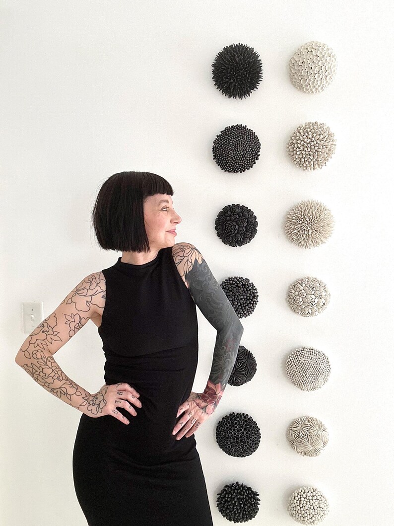 May include: A collection of decorative wall art pieces in various textures and colors, including black and off-white. The pieces are arranged in a grid pattern on a white wall. The art pieces have a round shape and a textured surface.