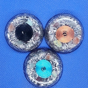 May include: Three round orgone energy generators with a silver matrix, each featuring a central spiral. One has a black spiral, another a copper spiral, and the third a turquoise spiral. Various small stones are embedded within the matrix.