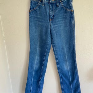 K&ouml;nnte beinhalten: Blaue Jeans aus Denim, die an einem transparenten Kleiderb&uuml;gel h&auml;ngen. Die Jeans haben ein klassisches F&uuml;nf-Taschen-Design, einen Knopf- und Rei&szlig;verschluss sowie kupferfarbene Nieten. Der Denim hat eine mittlere Waschung mit einigen Ausbleichungen und Gebrauchsspuren.