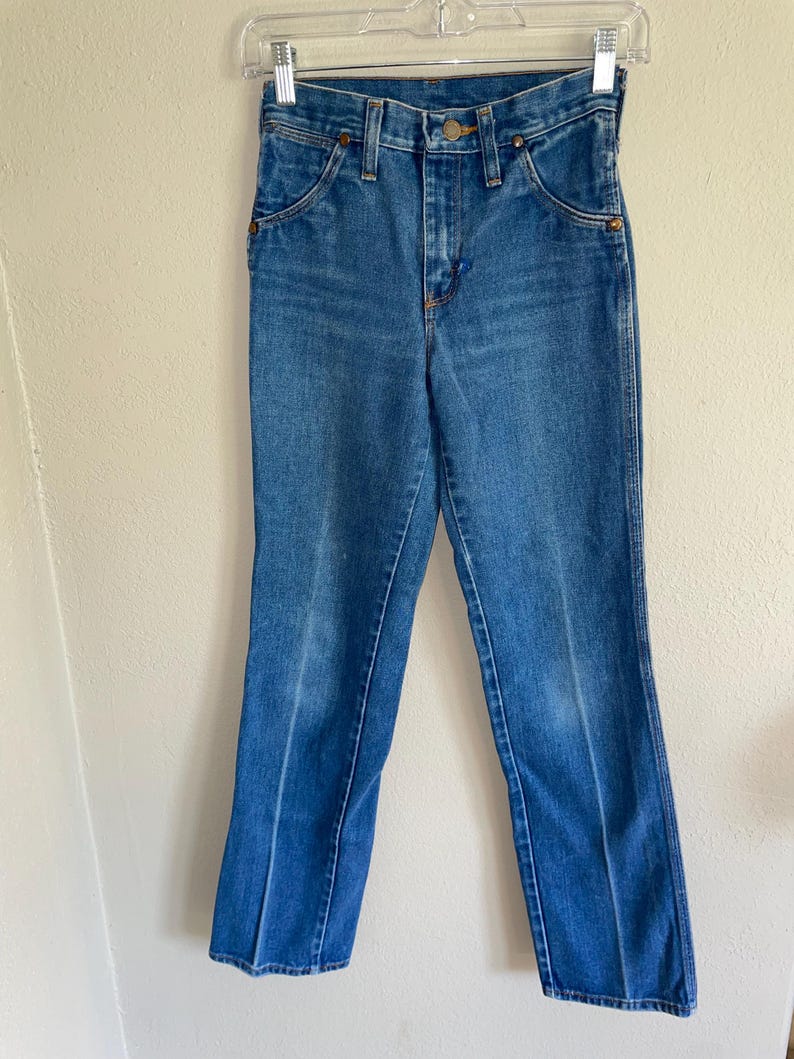 K&ouml;nnte beinhalten: Blaue Jeans aus Denim, die an einem transparenten Kleiderb&uuml;gel h&auml;ngen. Die Jeans haben ein klassisches F&uuml;nf-Taschen-Design, einen Knopf- und Rei&szlig;verschluss sowie kupferfarbene Nieten. Der Denim hat eine mittlere Waschung mit einigen Ausbleichungen und Gebrauchsspuren.