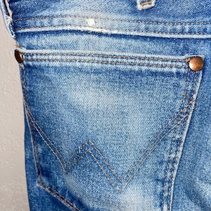 May include: Close-up of blue denim jeans, showing a back pocket with a distinctive W-shaped stitching. Copper rivets secure the pocket and waistband. The denim has a faded, worn appearance, typical of vintage jeans.
