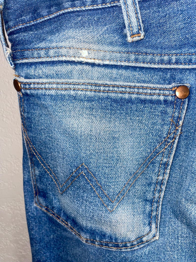 May include: Close-up of blue denim jeans, showing a back pocket with a distinctive W-shaped stitching. Copper rivets secure the pocket and waistband. The denim has a faded, worn appearance, typical of vintage jeans.