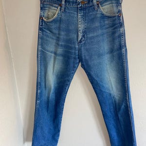 May include: Faded blue denim jeans with a classic five-pocket design. The jeans have a straight leg cut and show signs of wear, with light fading and distressing. They are hanging on a clear hanger.