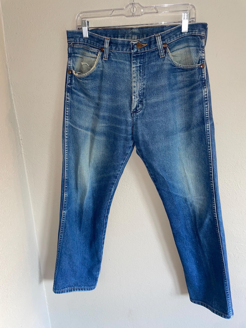 May include: Faded blue denim jeans with a classic five-pocket design. The jeans have a straight leg cut and show signs of wear, with light fading and distressing. They are hanging on a clear hanger.