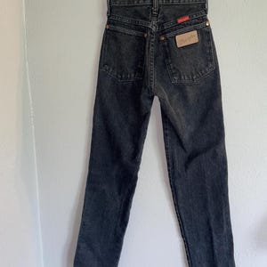 Op de afbeelding: Zwarte denim jeans hangend aan een houten hanger. De jeans hebben een hoge taille en een rechte pijp. Het Wrangler-merklabel is zichtbaar op de achterzak. De jeans hebben een vervaagde zwarte kleur.