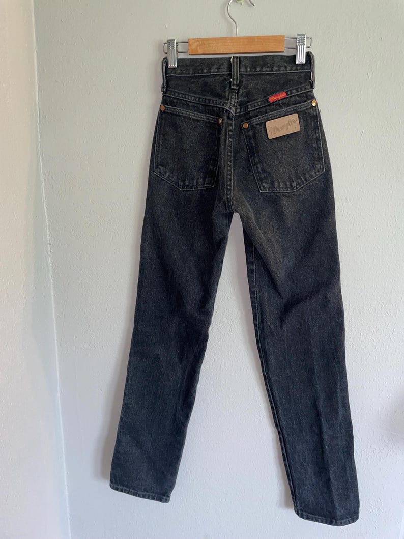 Op de afbeelding: Zwarte denim jeans hangend aan een houten hanger. De jeans hebben een hoge taille en een rechte pijp. Het Wrangler-merklabel is zichtbaar op de achterzak. De jeans hebben een vervaagde zwarte kleur.