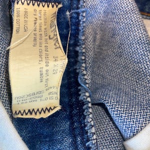 May include: Close-up of blue denim jeans with a white interior and a tag. The tag reads "Wrangler" and includes size 34 x 29, along with washing instructions and fabric content. The jeans are made of 100% cotton.