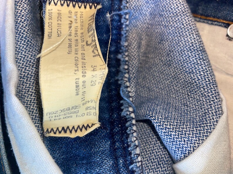 May include: Close-up of blue denim jeans with a white interior and a tag. The tag reads "Wrangler" and includes size 34 x 29, along with washing instructions and fabric content. The jeans are made of 100% cotton.