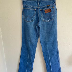 K&ouml;nnte beinhalten: Blaue Jeans aus Denim mit klassischer Passform, zwei Ges&auml;&szlig;taschen und einem braunen Lederpatch mit dem "Wrangler"-Logo. Die Jeans h&auml;ngen an einem transparenten Kunststoffb&uuml;gel.