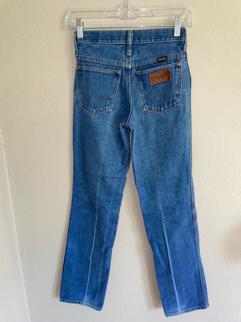 K&ouml;nnte beinhalten: Blaue Jeans aus Denim mit klassischer Passform, zwei Ges&auml;&szlig;taschen und einem braunen Lederpatch mit dem "Wrangler"-Logo. Die Jeans h&auml;ngen an einem transparenten Kunststoffb&uuml;gel.