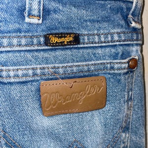 May include: Close-up of light blue denim jeans with the Wrangler brand label on the back pocket. The label is brown leather with the Wrangler logo embossed. A smaller black label with the Wrangler logo is above the back pocket.