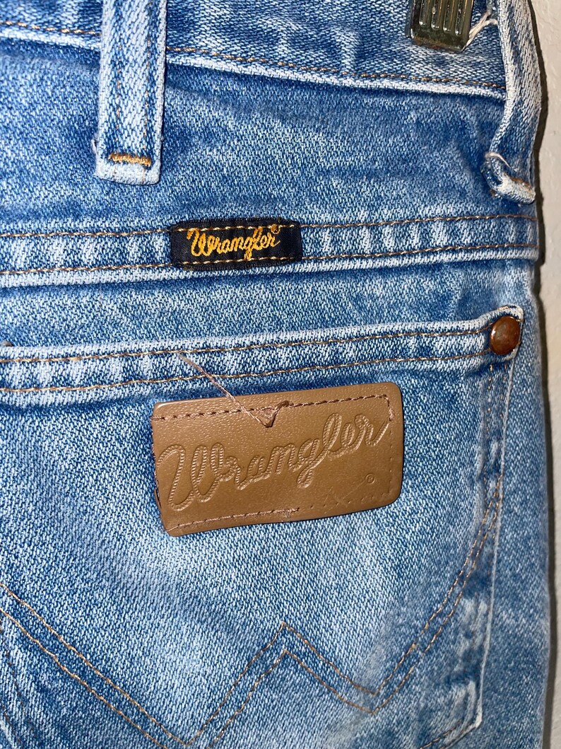 May include: Close-up of light blue denim jeans with the Wrangler brand label on the back pocket. The label is brown leather with the Wrangler logo embossed. A smaller black label with the Wrangler logo is above the back pocket.