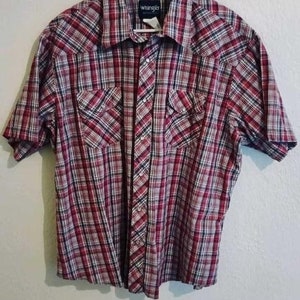 Puede incluir: Una camisa de manga corta a cuadros rojos, negros y blancos con una etiqueta de Wrangler.