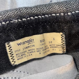 Op de afbeelding: Close-up van een Wrangler jeanslabel. Het cr&egrave;mekleurige label heeft zwarte tekst met maat 32 en wasinstructies. De jeans is van donkergrijze denim.