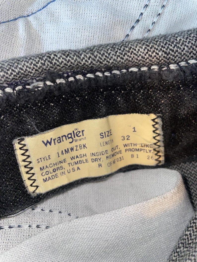 Op de afbeelding: Close-up van een Wrangler jeanslabel. Het cr&egrave;mekleurige label heeft zwarte tekst met maat 32 en wasinstructies. De jeans is van donkergrijze denim.