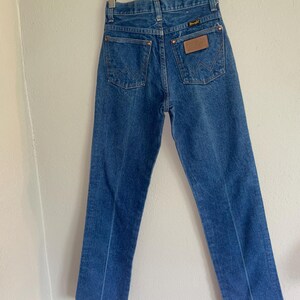 Puede incluir: Vaqueros de mezclilla azules colgados de una percha de madera. Los vaqueros tienen un dise&ntilde;o cl&aacute;sico de cinco bolsillos con el logotipo de Wrangler en el bolsillo trasero. Los vaqueros son de un lavado medio y parecen ser vintage.