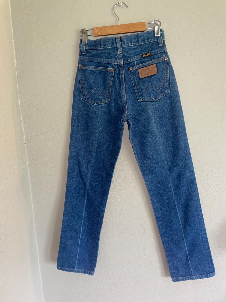 Puede incluir: Vaqueros de mezclilla azules colgados de una percha de madera. Los vaqueros tienen un dise&ntilde;o cl&aacute;sico de cinco bolsillos con el logotipo de Wrangler en el bolsillo trasero. Los vaqueros son de un lavado medio y parecen ser vintage.