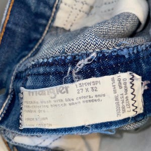 K&ouml;nnte beinhalten: Nahaufnahme einer blauen Jeans mit einem Wrangler-Etikett. Das Etikett gibt die Gr&ouml;&szlig;e von 68,58 x 81,28 cm an. Die Jeans besteht aus 100 % Baumwolle und enth&auml;lt Waschanweisungen. Die Jeans wird in den USA hergestellt.