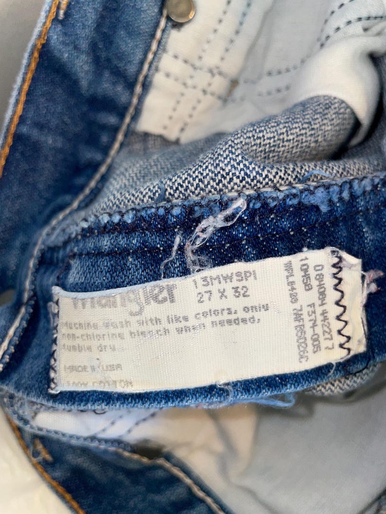 K&ouml;nnte beinhalten: Nahaufnahme einer blauen Jeans mit einem Wrangler-Etikett. Das Etikett gibt die Gr&ouml;&szlig;e von 68,58 x 81,28 cm an. Die Jeans besteht aus 100 % Baumwolle und enth&auml;lt Waschanweisungen. Die Jeans wird in den USA hergestellt.
