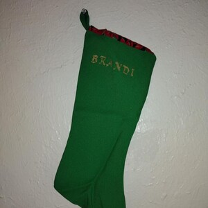 May include: Green Christmas stocking with a red and black floral pattern at the top. The stocking is personalized with the name "Brandi" in gold thread.