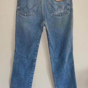 May include: Light blue denim jeans with a classic five-pocket design. The jeans feature a straight leg cut and a brown leather patch with the brand name. The jeans are hanging on a wooden hanger.