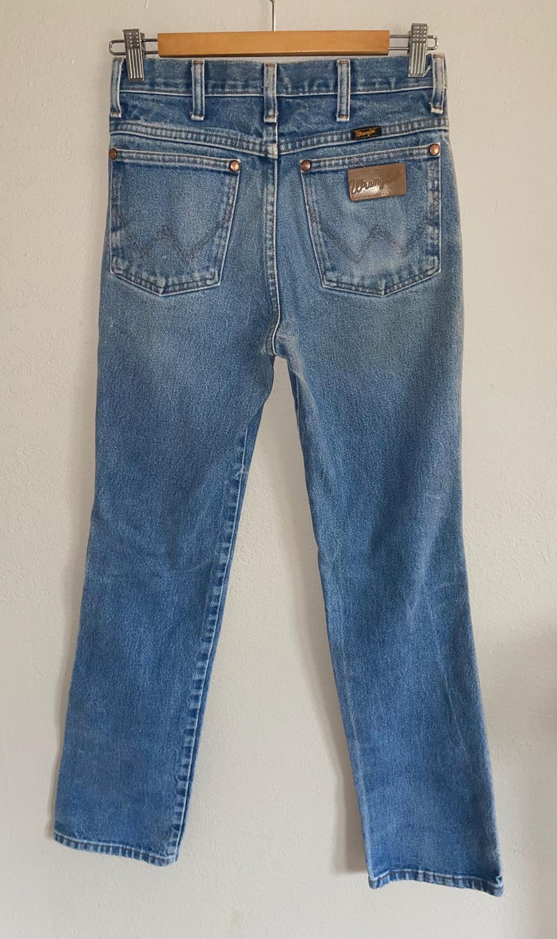 May include: Light blue denim jeans with a classic five-pocket design. The jeans feature a straight leg cut and a brown leather patch with the brand name. The jeans are hanging on a wooden hanger.