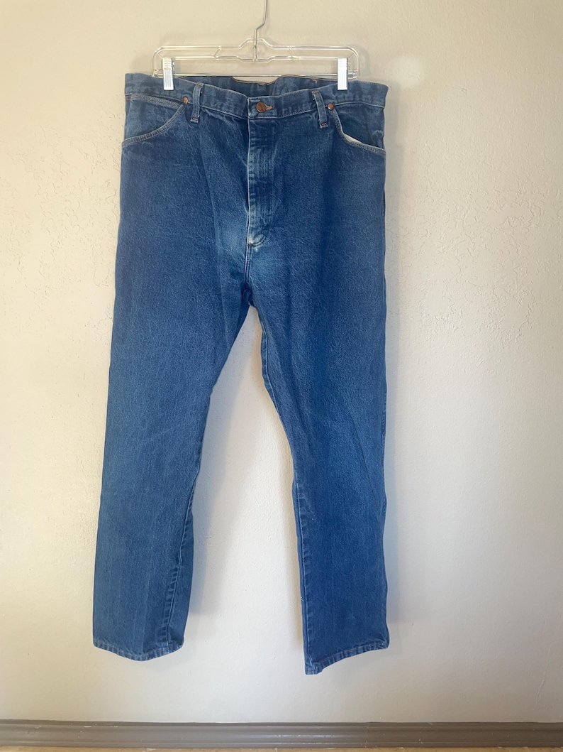 May include: A pair of blue denim jeans hanging on a coat hanger. The jeans have a classic straight leg style and a button fly.