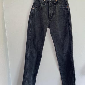 Op de afbeelding: Een zwarte denim jeans hangend aan een houten hanger. De jeans heeft een hoge taille, een knoopsluiting en een klassiek vijf-zakken ontwerp. De denim heeft een vervaagde, versleten uitstraling.
