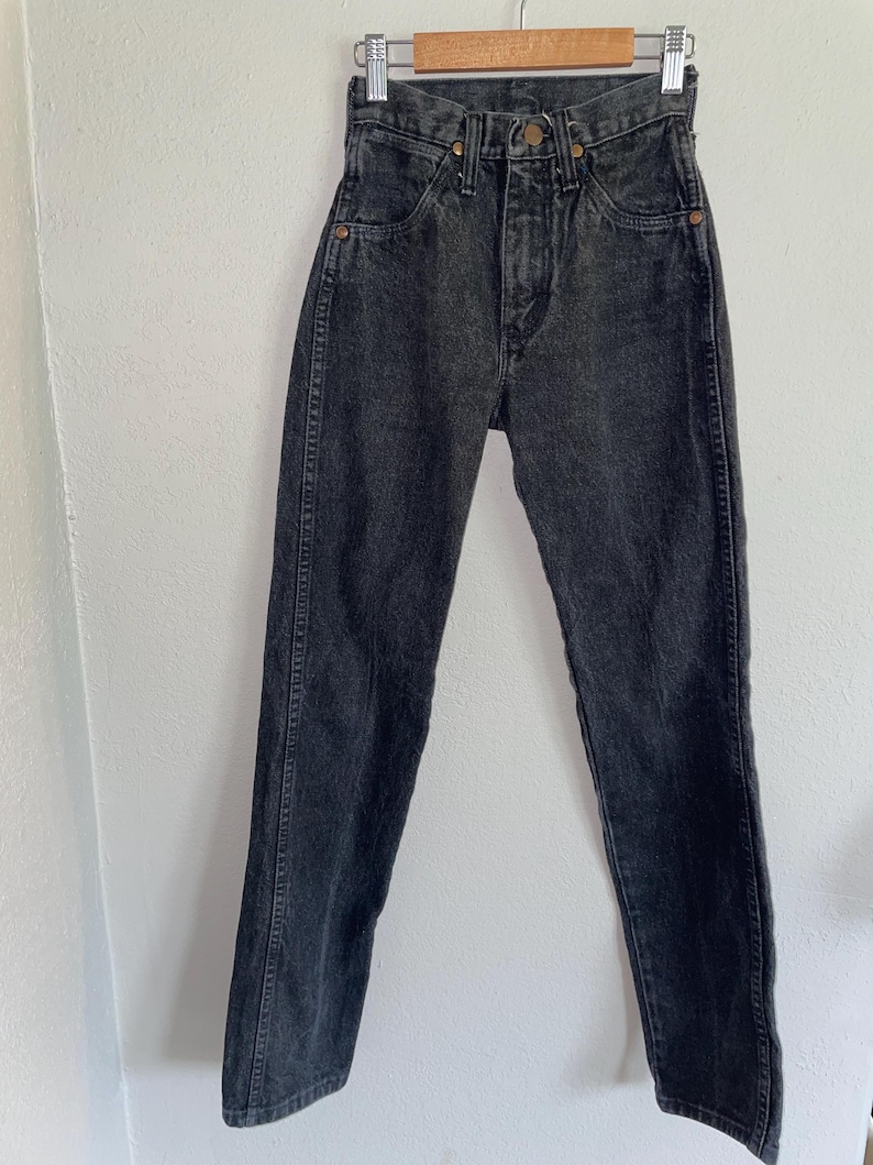 Op de afbeelding: Een zwarte denim jeans hangend aan een houten hanger. De jeans heeft een hoge taille, een knoopsluiting en een klassiek vijf-zakken ontwerp. De denim heeft een vervaagde, versleten uitstraling.