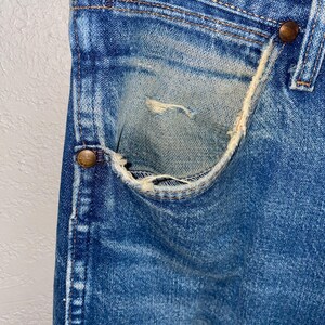 May include: Close-up of vintage blue denim jeans, showing a worn pocket with distressed details. The jeans have copper-coloured rivets and visible stitching. The denim has faded areas, adding to the vintage aesthetic.