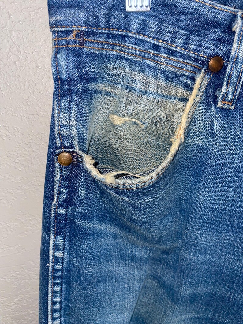 May include: Close-up of vintage blue denim jeans, showing a worn pocket with distressed details. The jeans have copper-coloured rivets and visible stitching. The denim has faded areas, adding to the vintage aesthetic.