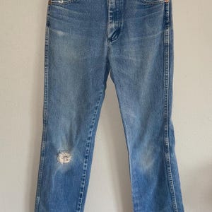 May include: Light blue denim jeans with a high waist and a distressed knee. The jeans have a classic five-pocket design and a metal zipper and button closure. The jeans are hanging on a metal hanger.