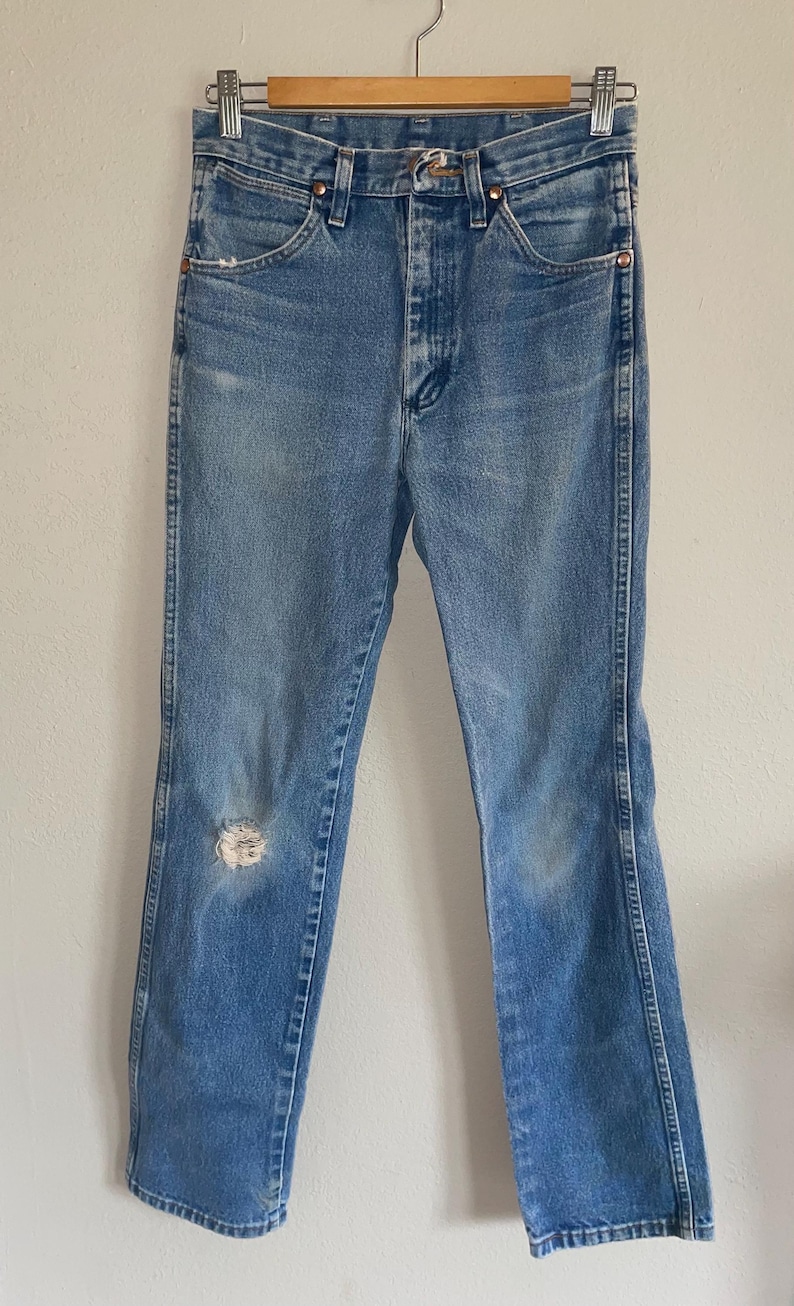 May include: Light blue denim jeans with a high waist and a distressed knee. The jeans have a classic five-pocket design and a metal zipper and button closure. The jeans are hanging on a metal hanger.