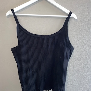 May include: A black ribbed tank top with a scoop neck and thin straps. The top has a slightly cropped length and a ruffled hem.