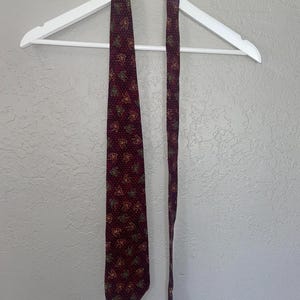 May include: A burgundy silk tie with a repeating pattern of small, colorful figures. The tie is hanging on a white hanger.