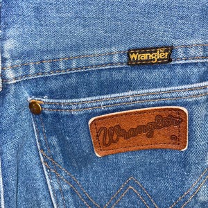 K&ouml;nnte beinhalten: Nahaufnahme einer blauen Jeans mit einem braunen Leder-Wrangler-Patch auf der Ges&auml;&szlig;tasche. Die Jeans hat ein klassisches Design mit sichtbaren N&auml;hten und einem kleinen, goldfarbenen Knopf.