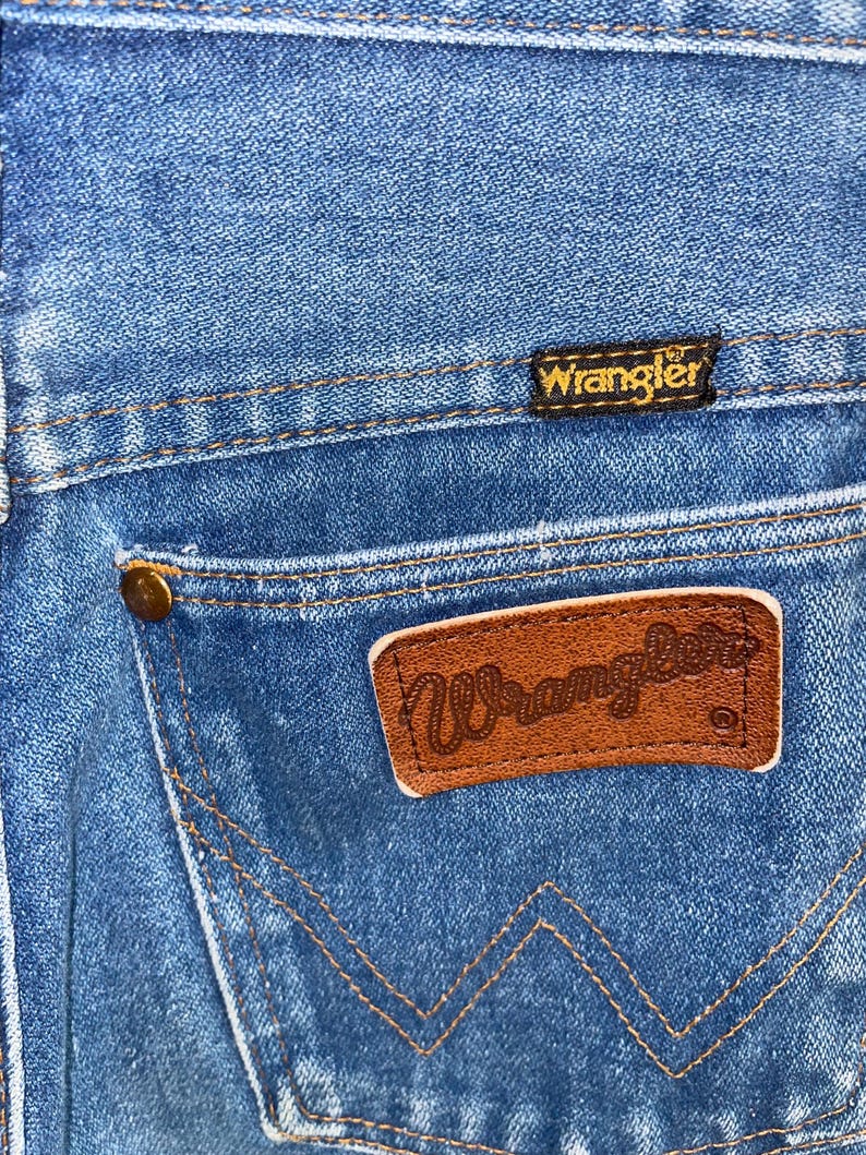 K&ouml;nnte beinhalten: Nahaufnahme einer blauen Jeans mit einem braunen Leder-Wrangler-Patch auf der Ges&auml;&szlig;tasche. Die Jeans hat ein klassisches Design mit sichtbaren N&auml;hten und einem kleinen, goldfarbenen Knopf.