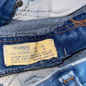 K&ouml;nnte beinhalten: Nahaufnahme einer blauen Wrangler-Jeans. Die Jeans hat ein gelbes Etikett mit dem Markennamen und Waschanweisungen. Die Bundweite betr&auml;gt 66 cm und die L&auml;nge 81 cm. Die Jeans wird in den USA hergestellt.