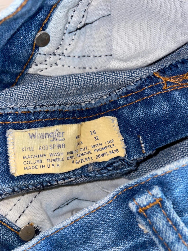 K&ouml;nnte beinhalten: Nahaufnahme einer blauen Wrangler-Jeans. Die Jeans hat ein gelbes Etikett mit dem Markennamen und Waschanweisungen. Die Bundweite betr&auml;gt 66 cm und die L&auml;nge 81 cm. Die Jeans wird in den USA hergestellt.