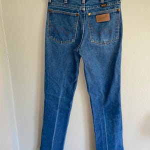 K&ouml;nnte beinhalten: Blaue Jeans aus Denim mit dem Wrangler-Logo auf der Ges&auml;&szlig;tasche. Die Jeans haben ein klassisches F&uuml;nf-Taschen-Design mit kupferfarbenen Nieten und N&auml;hten. Die Jeans h&auml;ngen an einem Kleiderb&uuml;gel.