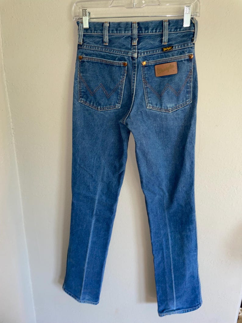 K&ouml;nnte beinhalten: Blaue Jeans aus Denim mit dem Wrangler-Logo auf der Ges&auml;&szlig;tasche. Die Jeans haben ein klassisches F&uuml;nf-Taschen-Design mit kupferfarbenen Nieten und N&auml;hten. Die Jeans h&auml;ngen an einem Kleiderb&uuml;gel.