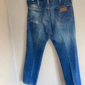 May include: Blue denim jeans with a classic five-pocket design. The jeans have a faded wash and a leather patch with the brand name on the back pocket. The jeans are hanging on a clear plastic hanger.