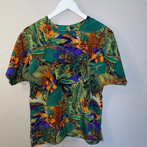 May include: A short-sleeved, green, purple, orange and brown floral print blouse with a round neckline. The blouse is hanging on a white hanger.
