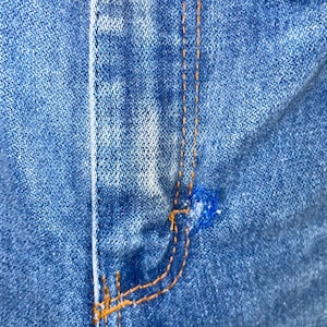 K&ouml;nnte beinhalten: Nahaufnahme von blauem Denim-Stoff mit orangefarbenen N&auml;hten. Das Bild zeigt die Naht einer Tasche und den Hosenschlitz einer Jeans. Der Denim hat ein abgenutztes, verwaschenes Aussehen.