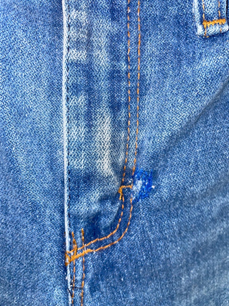 K&ouml;nnte beinhalten: Nahaufnahme von blauem Denim-Stoff mit orangefarbenen N&auml;hten. Das Bild zeigt die Naht einer Tasche und den Hosenschlitz einer Jeans. Der Denim hat ein abgenutztes, verwaschenes Aussehen.