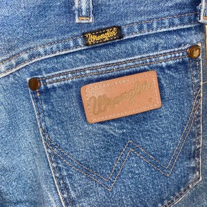 May include: Close-up of blue denim jeans showing a back pocket with the Wrangler logo. The pocket has a brown leather patch with the Wrangler brand name embossed. The jeans have a classic five-pocket design with copper-coloured rivets.
