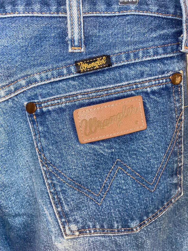 May include: Close-up of blue denim jeans showing a back pocket with the Wrangler logo. The pocket has a brown leather patch with the Wrangler brand name embossed. The jeans have a classic five-pocket design with copper-coloured rivets.
