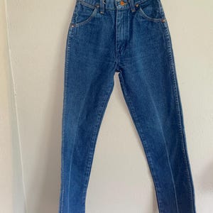 Puede incluir: Vaqueros de mezclilla azules colgados de una percha de madera. Los vaqueros tienen una cintura alta, un dise&ntilde;o cl&aacute;sico de cinco bolsillos y una pernera recta. Los vaqueros son de un lavado medio con costuras visibles y remaches de cobre.