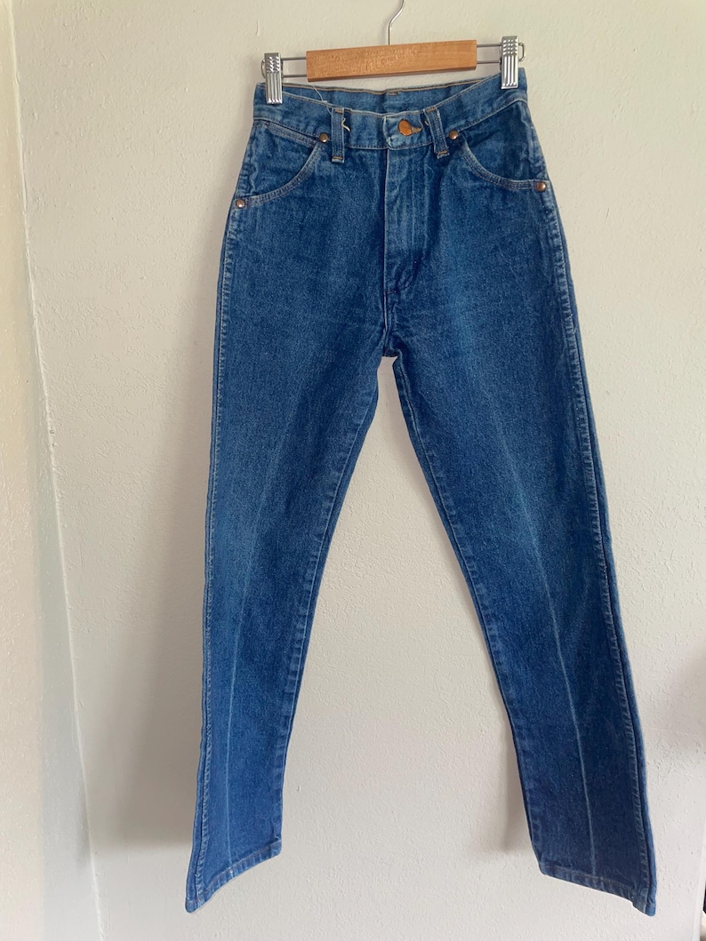 Puede incluir: Vaqueros de mezclilla azules colgados de una percha de madera. Los vaqueros tienen una cintura alta, un dise&ntilde;o cl&aacute;sico de cinco bolsillos y una pernera recta. Los vaqueros son de un lavado medio con costuras visibles y remaches de cobre.